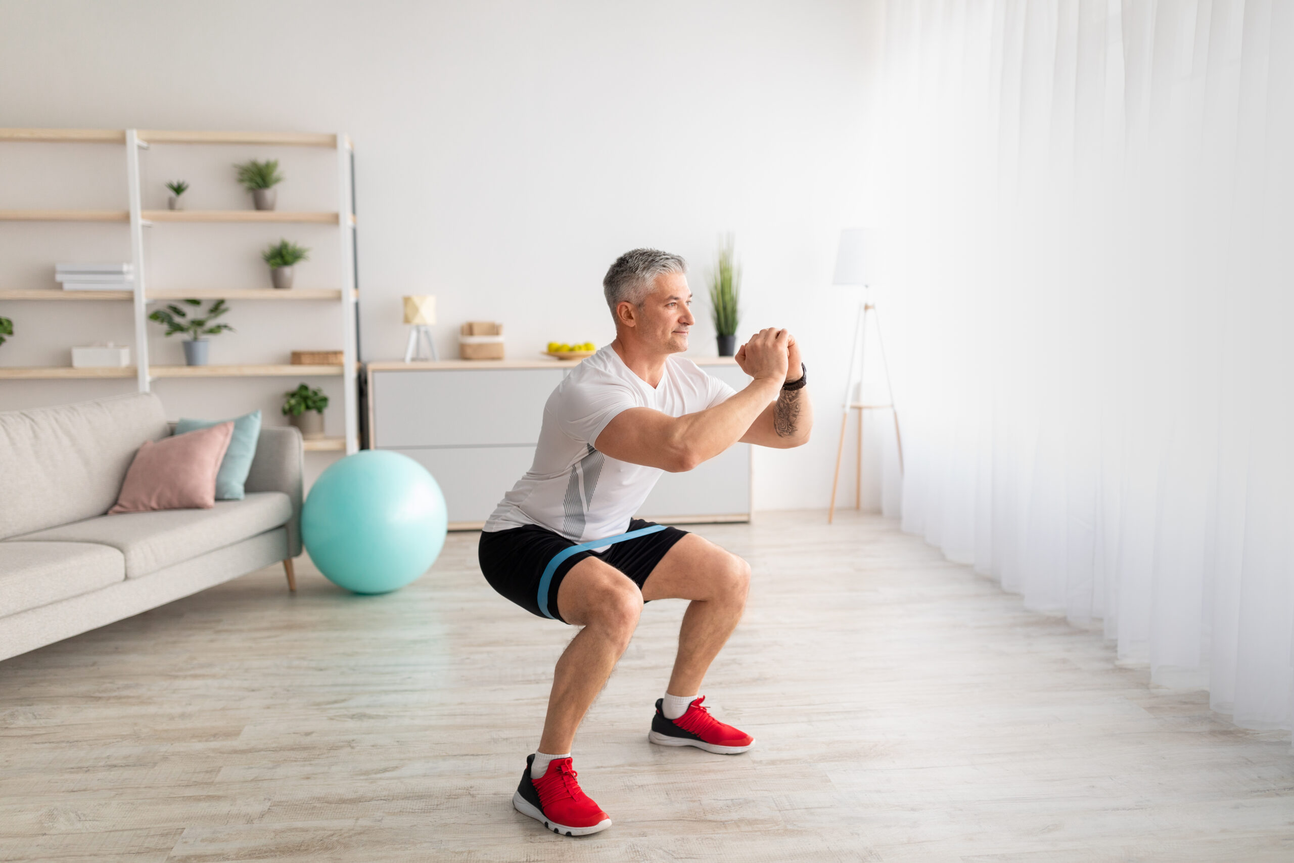 Morning training. Sporty mature man doing squats, exercising in living room interior, copy space. Athletic sportsman having domestic training during covid-19 lockdown